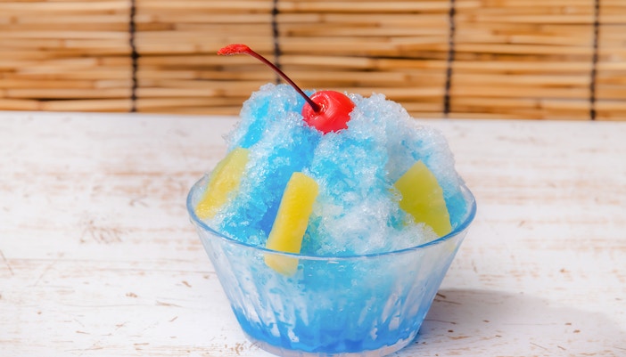 Shaved ice with blue syrup, pineapple slices, and a cherry on top in a glass bowl.
