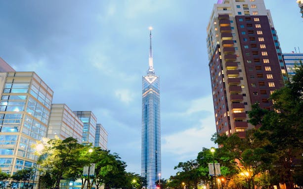 Fukuoka Tower illuminated at dusk in Fukuoka City, surrounded by modern buildings.