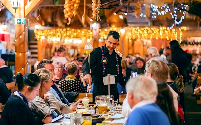 Server pouring drinks at a Prague folklore dinner show with guests seated at tables.