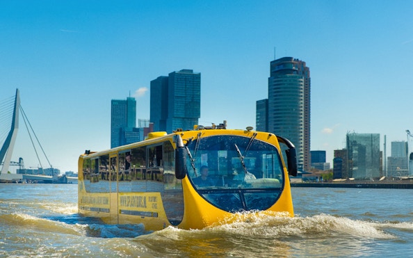 Amphibious bus on Rotterdam waterway with city skyline in background.