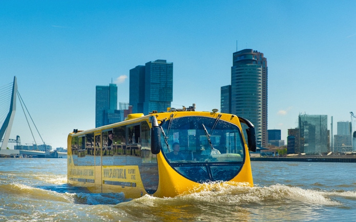 Amphibious bus on Rotterdam waterway with city skyline in background.