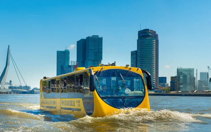 Amphibious bus on Rotterdam waterway with city skyline in background.