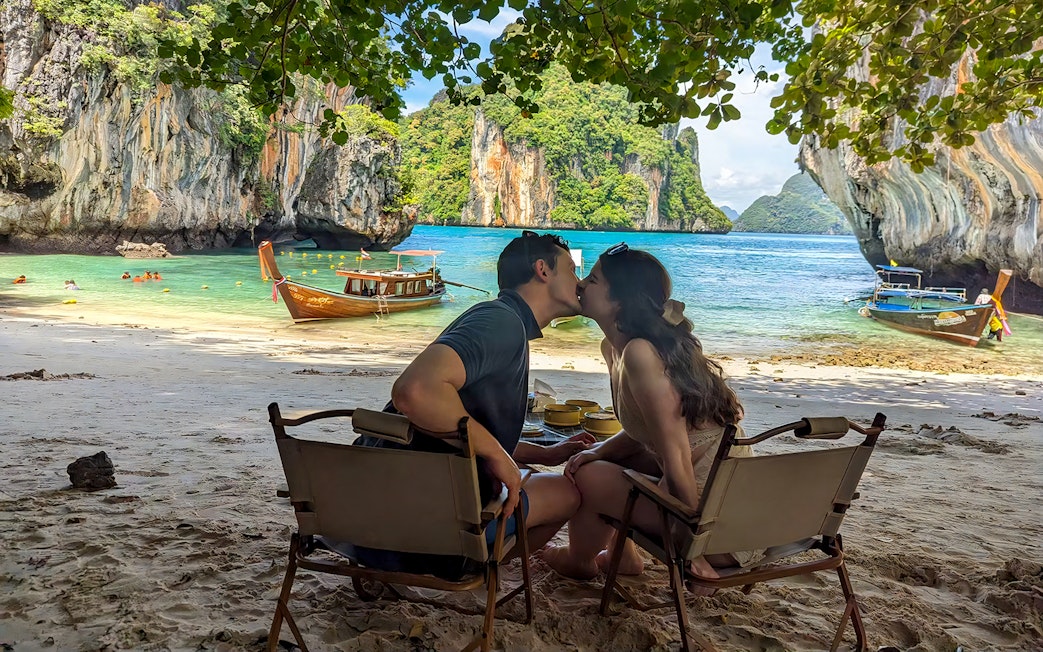 Couple sitting on Krabi Hong Island beach with speed boats and limestone cliffs in the background.