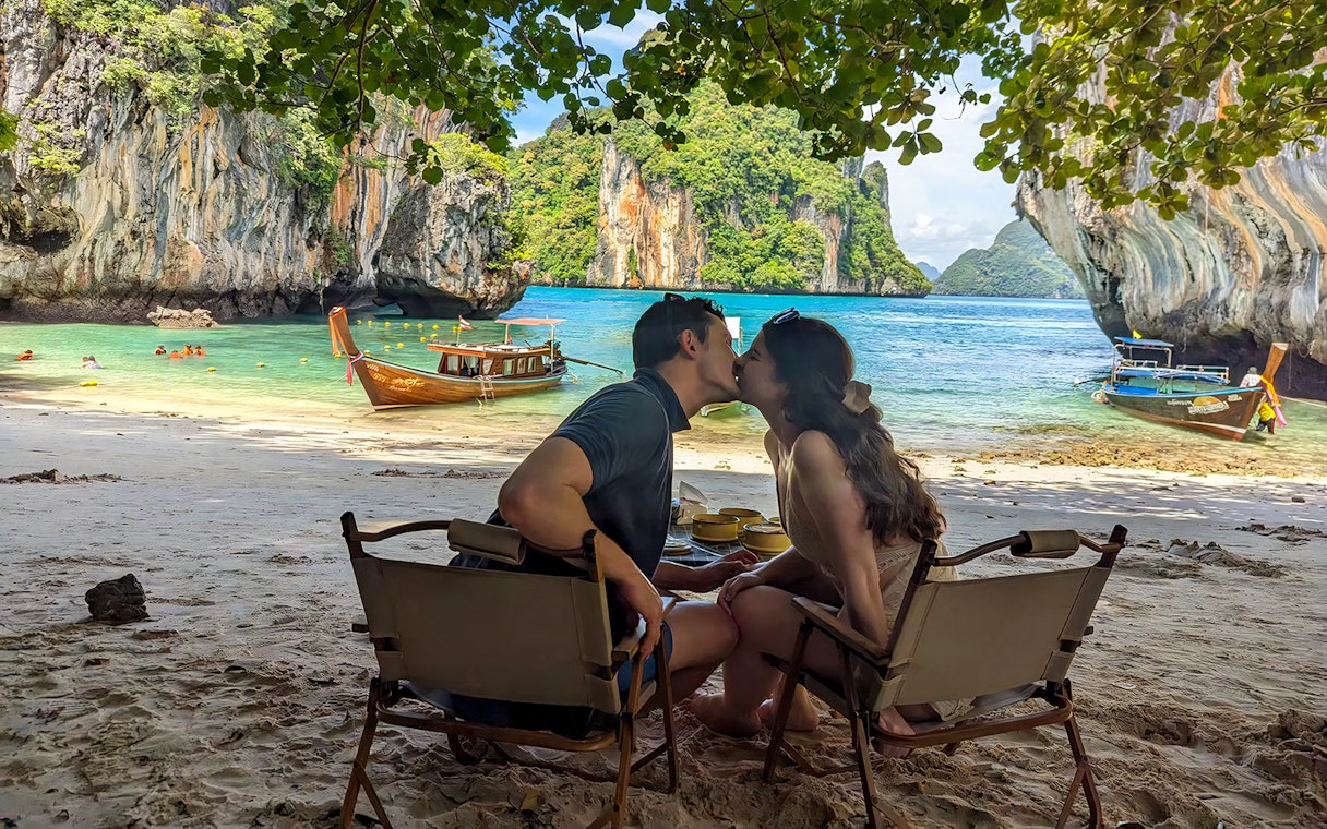 Couple sitting on Krabi Hong Island beach with speed boats and limestone cliffs in the background.