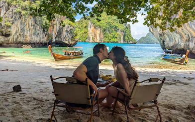 Couple sitting on Krabi Hong Island beach with speed boats and limestone cliffs in the background.