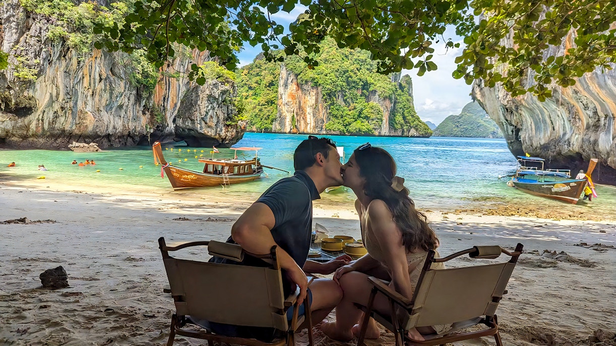 Couple sitting on Krabi Hong Island beach with speed boats and limestone cliffs in the background.