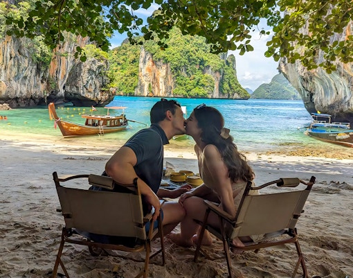 Couple sitting on Krabi Hong Island beach with speed boats and limestone cliffs in the background.