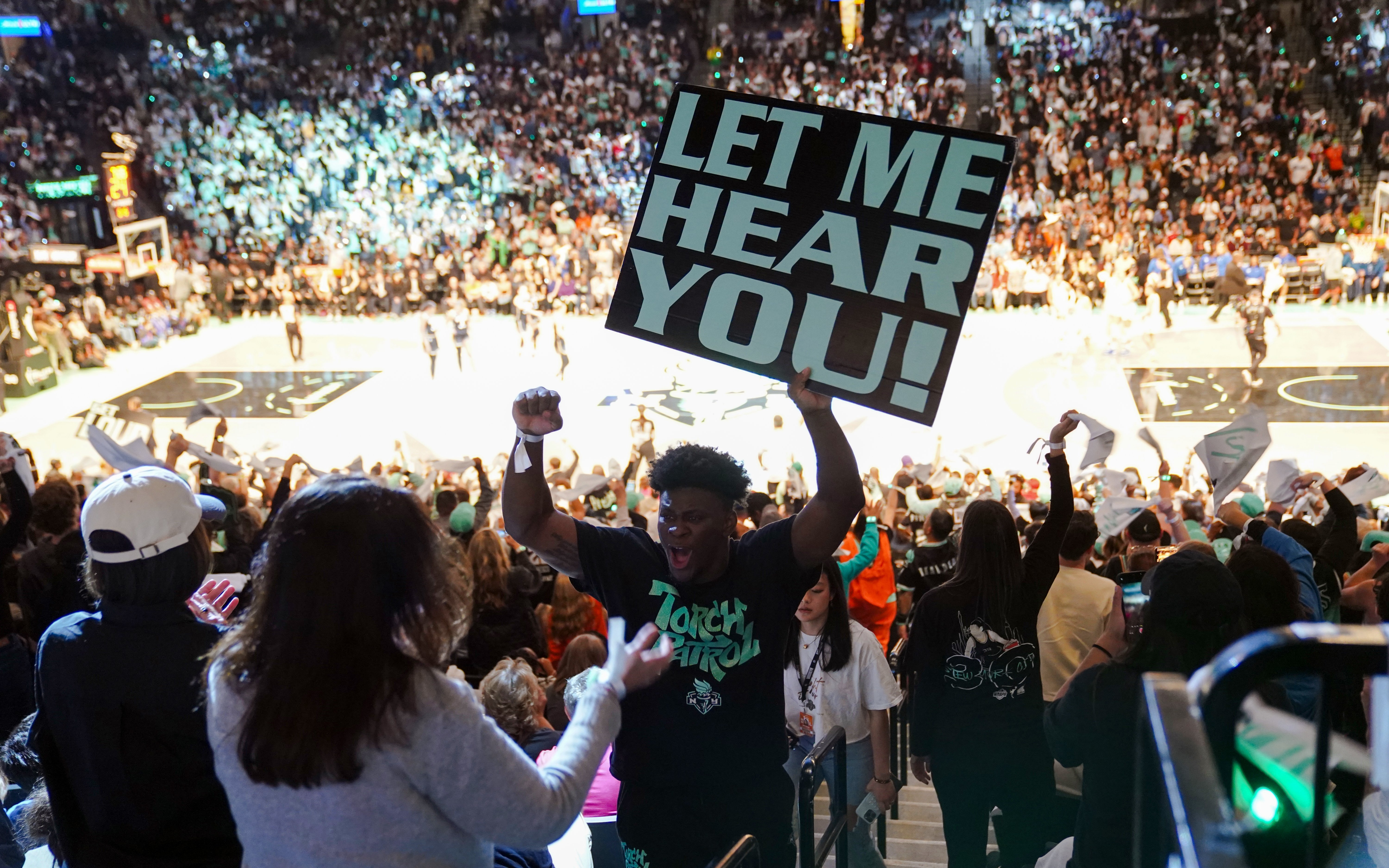 Fans cheering at New York Liberty basketball game with a "Let Me Hear You" sign.