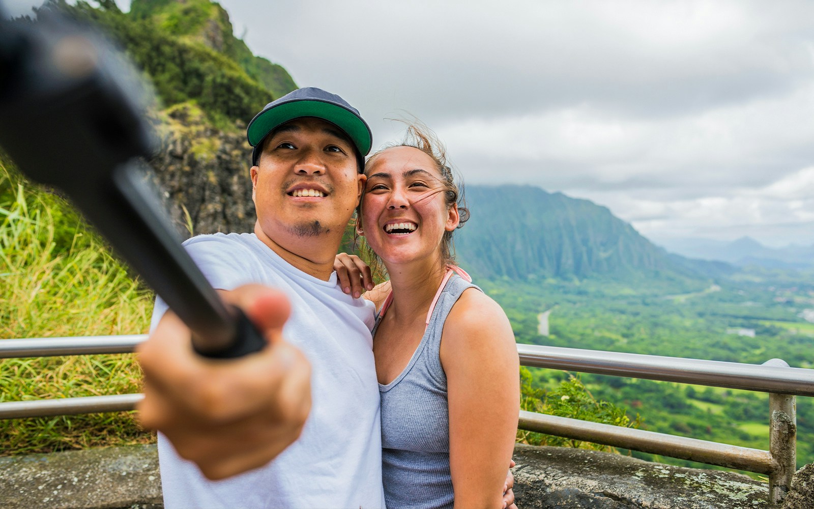 Couple taking selfie at Nuuanu Pali Lookout with lush mountains in the background.