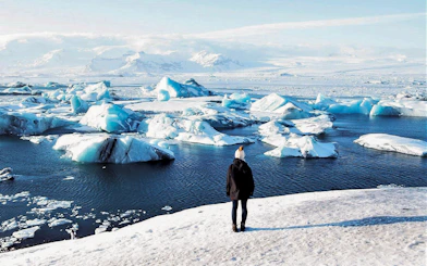 Person standing on snowy shore overlooking icebergs in Jökulsárlón Glacier Lagoon, South Iceland.