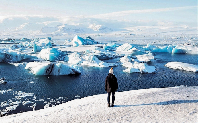 Person standing on snowy shore overlooking icebergs in Jökulsárlón Glacier Lagoon, South Iceland.