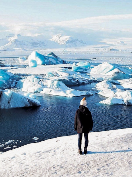 Person standing on snowy shore overlooking icebergs in Jökulsárlón Glacier Lagoon, South Iceland.