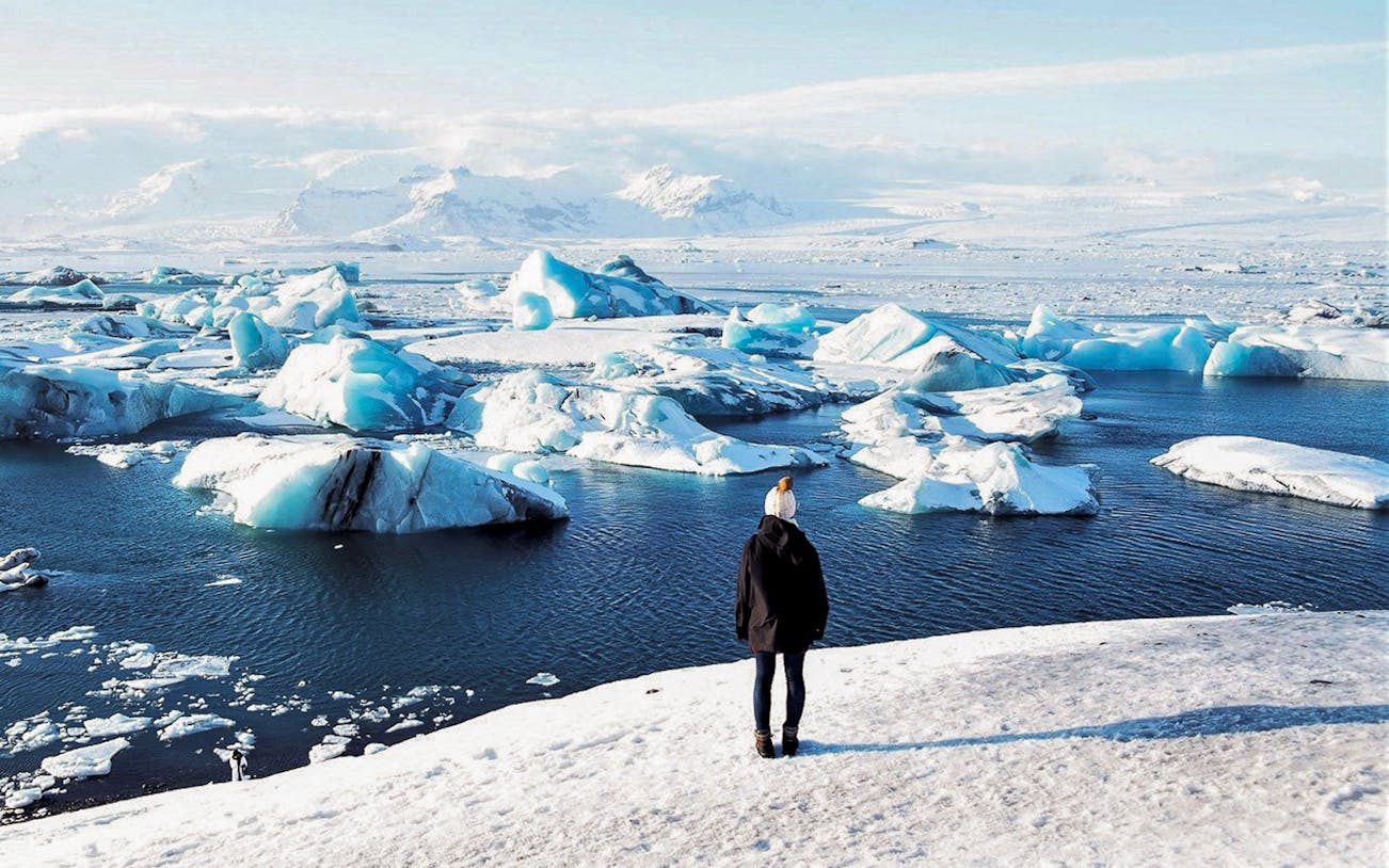 Person standing on snowy shore overlooking icebergs in Jökulsárlón Glacier Lagoon, South Iceland.