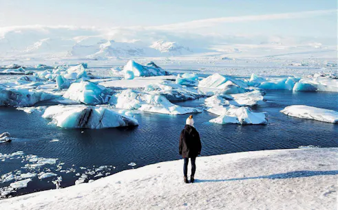 Person standing on snowy shore overlooking icebergs in Jökulsárlón Glacier Lagoon, South Iceland.