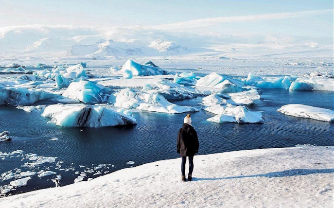 Person standing on snowy shore overlooking icebergs in Jökulsárlón Glacier Lagoon, South Iceland.