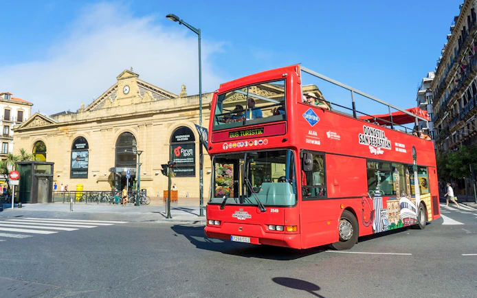 Red double-decker bus on San Sebastian city tour near historic building.