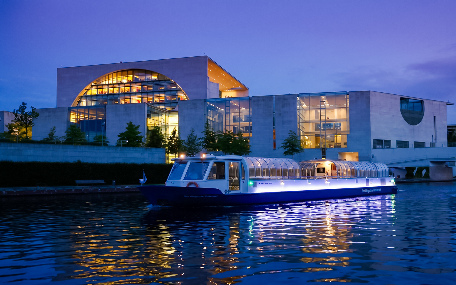 Dinner cruise boat on Berlin's Spree River at dusk near modern glass building.