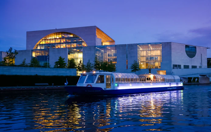 Dinner cruise boat on Berlin's Spree River at dusk near modern glass building.