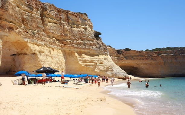 Beachgoers enjoy a sunny day near Benagil Caves, Algarve, with BBQ setup and umbrellas.