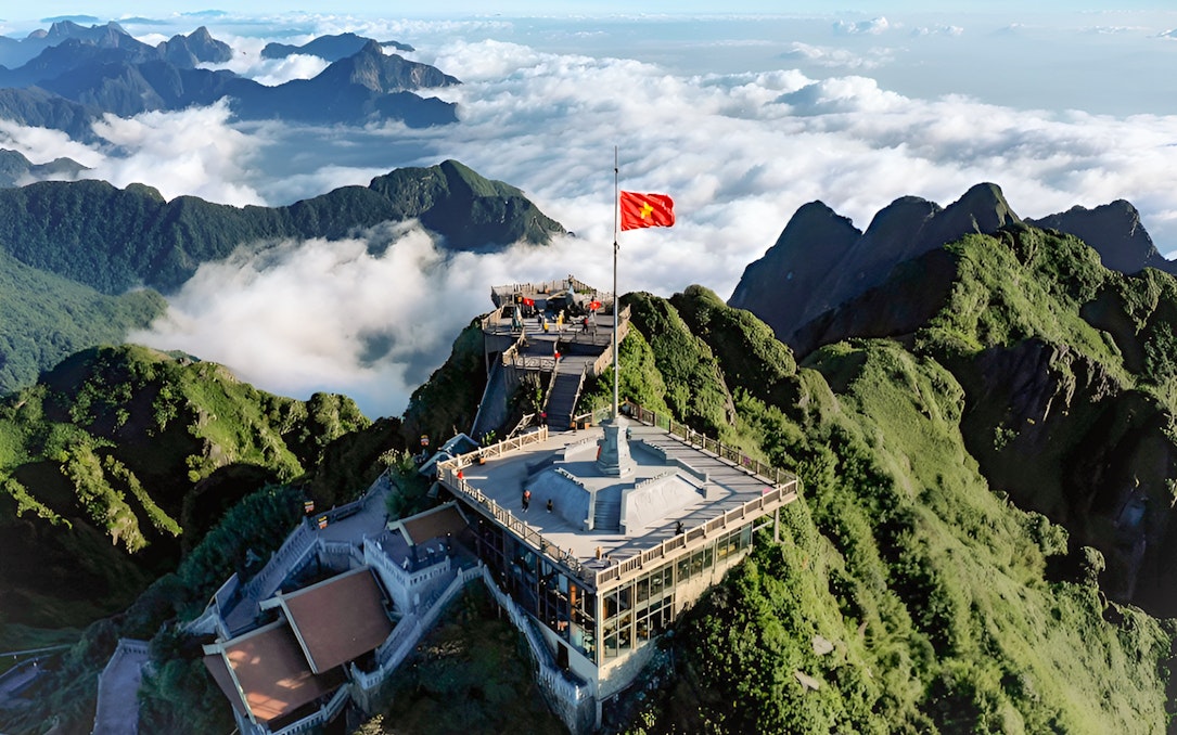 Sun World Fansipan Legend summit with flag, surrounded by lush mountains and clouds in Vietnam.