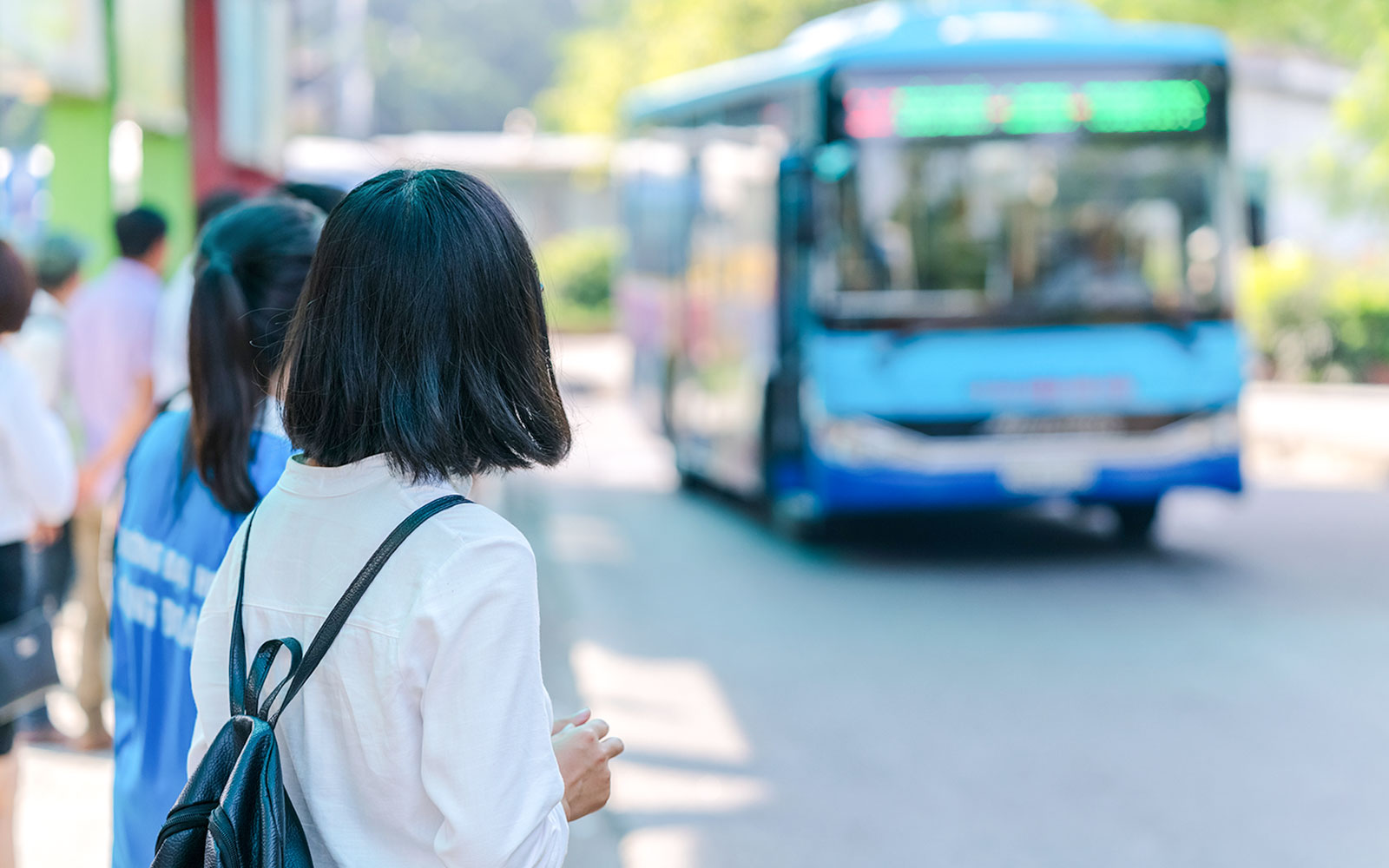 Young girl with backpack waiting at bus station as blue bus approaches.