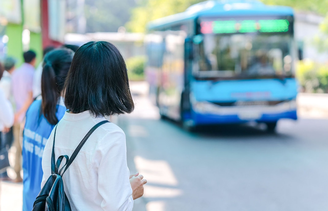 Young girl with backpack waiting at bus station as blue bus approaches.