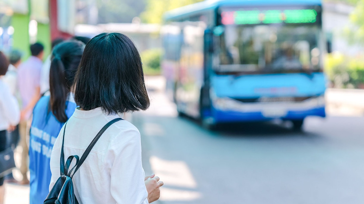 Young girl with backpack waiting at bus station as blue bus approaches.