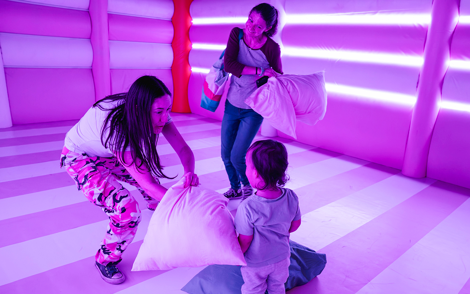 Adults and child playing with pillows in a vibrant, neon-lit room at Dopamine Land's Cushion Clash.
