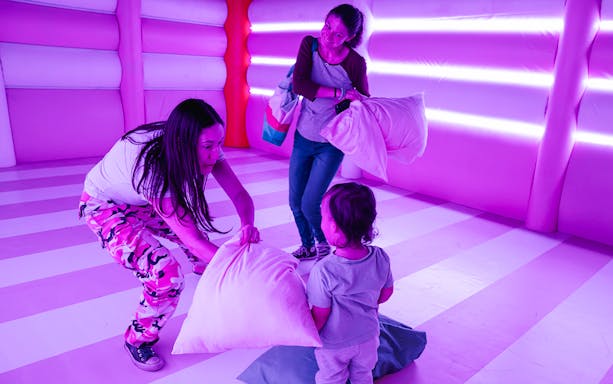 Adults and child playing with pillows in a vibrant, neon-lit room at Dopamine Land's Cushion Clash.