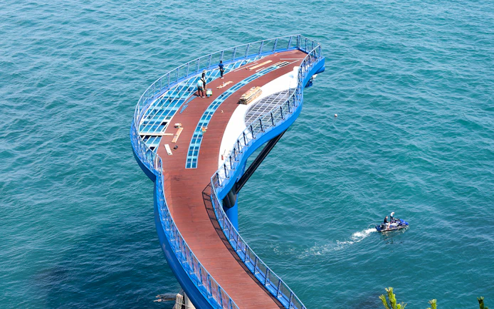 Blueline Park Sky Capsule platform over ocean in Busan, South Korea, with people and jet ski nearby.