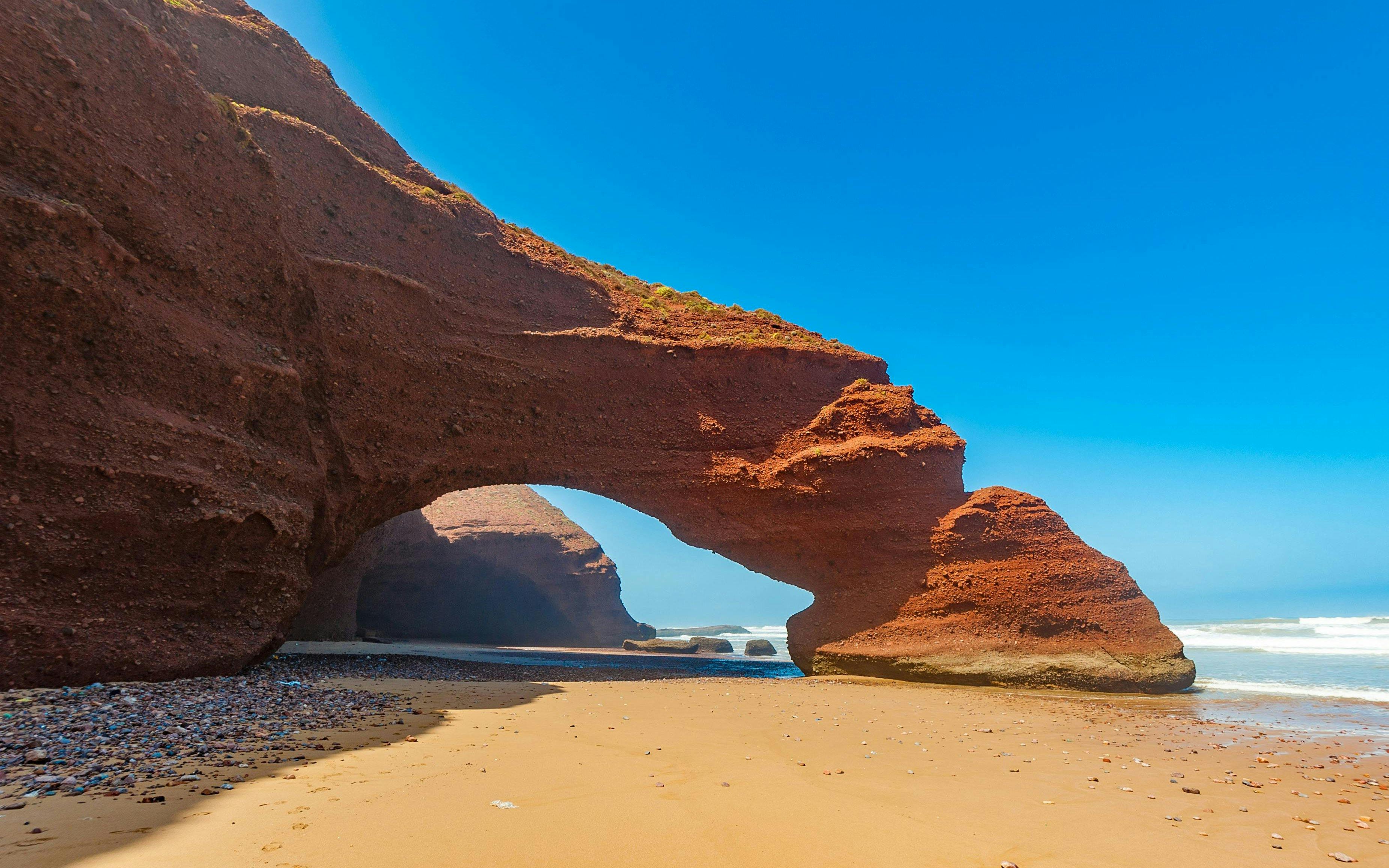 Red rock arch formations on Legzira Beach, Morocco, with ocean waves in the background.