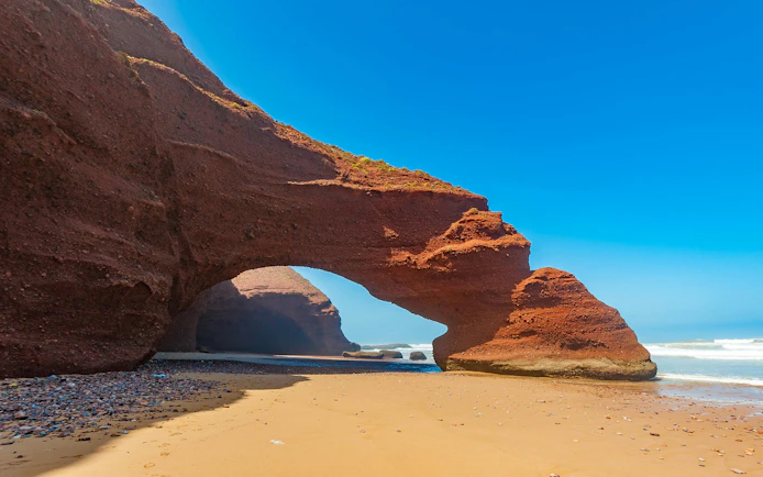 Red rock arch formations on Legzira Beach, Morocco, with ocean waves in the background.