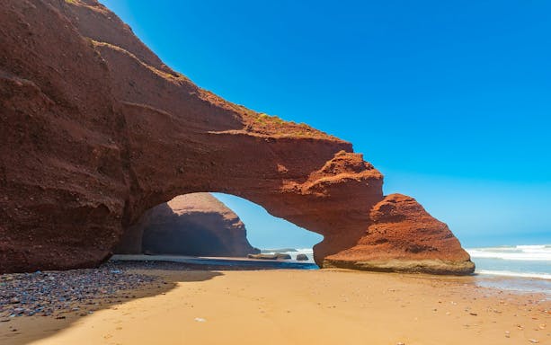 Red rock arch formations on Legzira Beach, Morocco, with ocean waves in the background.