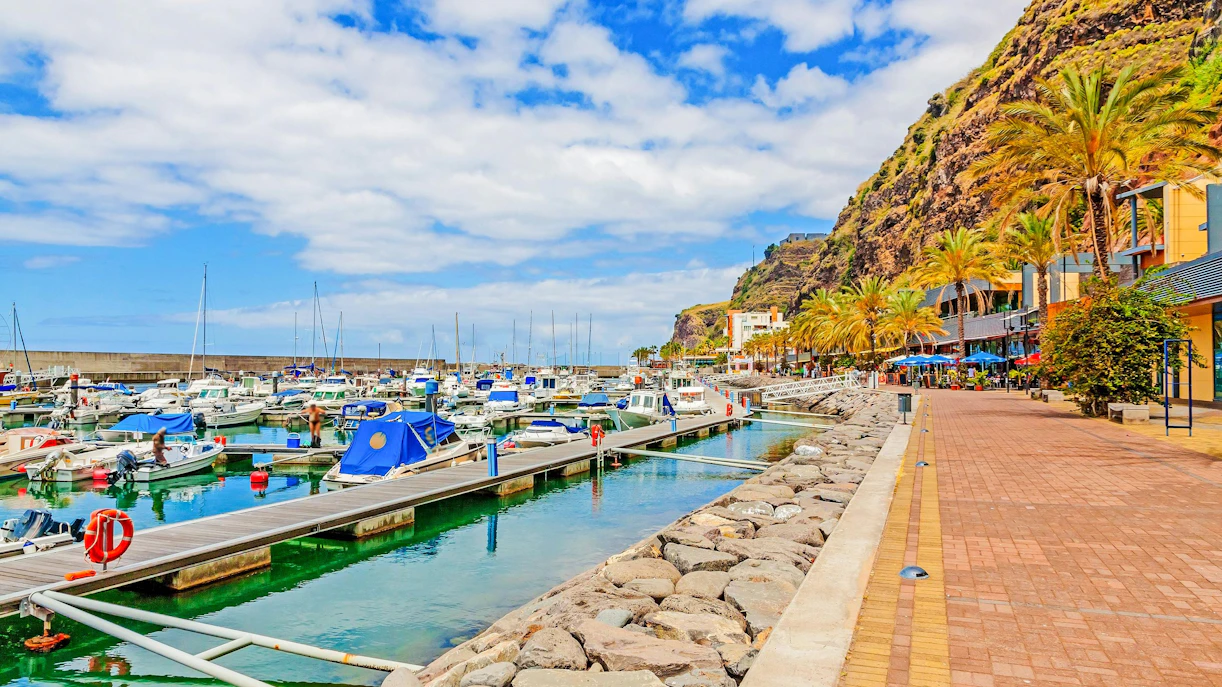 Boats docked at Calheta port, Madeira, with a promenade lined by palm trees and cliffs in the background.