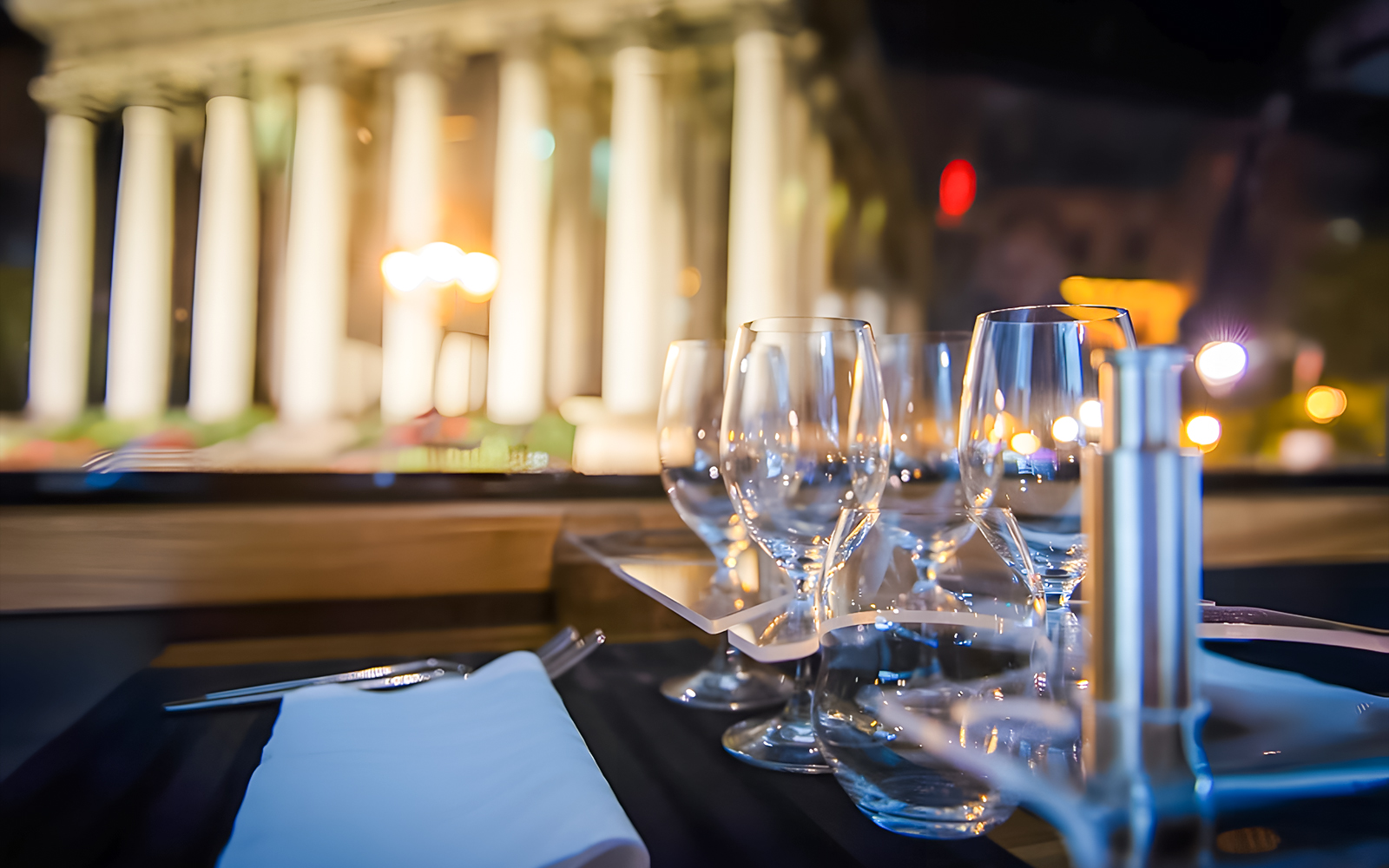 Cutlery and wine glasses on a table inside Bustronome Paris with blurred city lights.