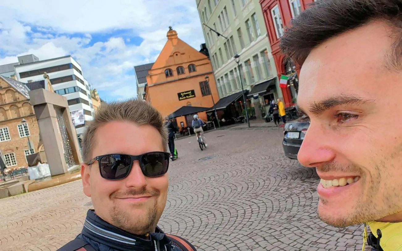 Two people smiling in a cobblestone square in Oslo, with historic buildings in the background.
