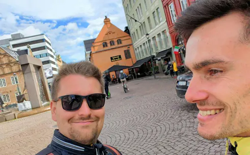 Two people smiling in a cobblestone square in Oslo, with historic buildings in the background.