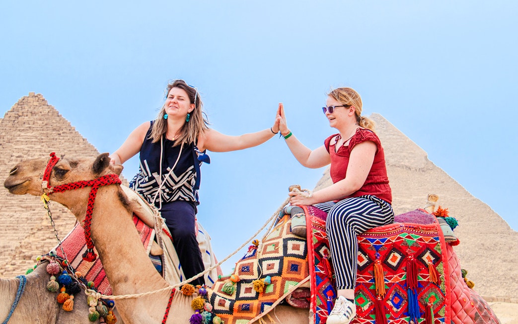 Two women high-fiving while riding camels near the Pyramids of Giza, Egypt.