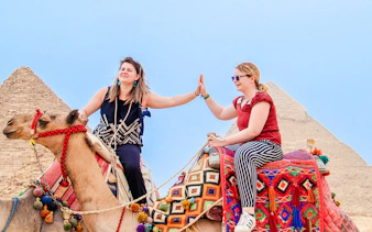 Two women high-fiving while riding camels near the Pyramids of Giza, Egypt.