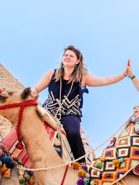 Two women high-fiving while riding camels near the Pyramids of Giza, Egypt.