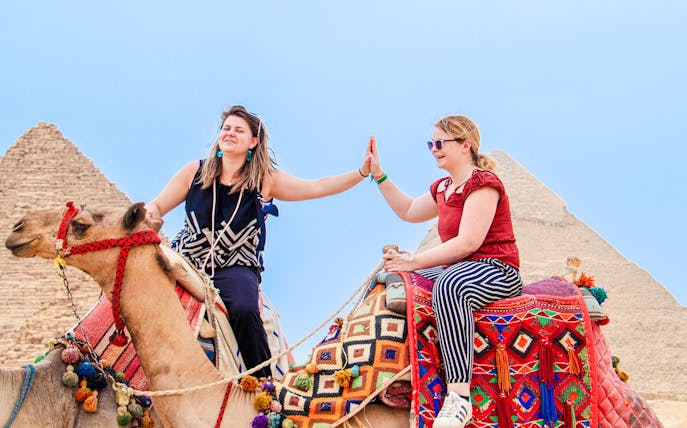 Two women high-fiving while riding camels near the Pyramids of Giza, Egypt.
