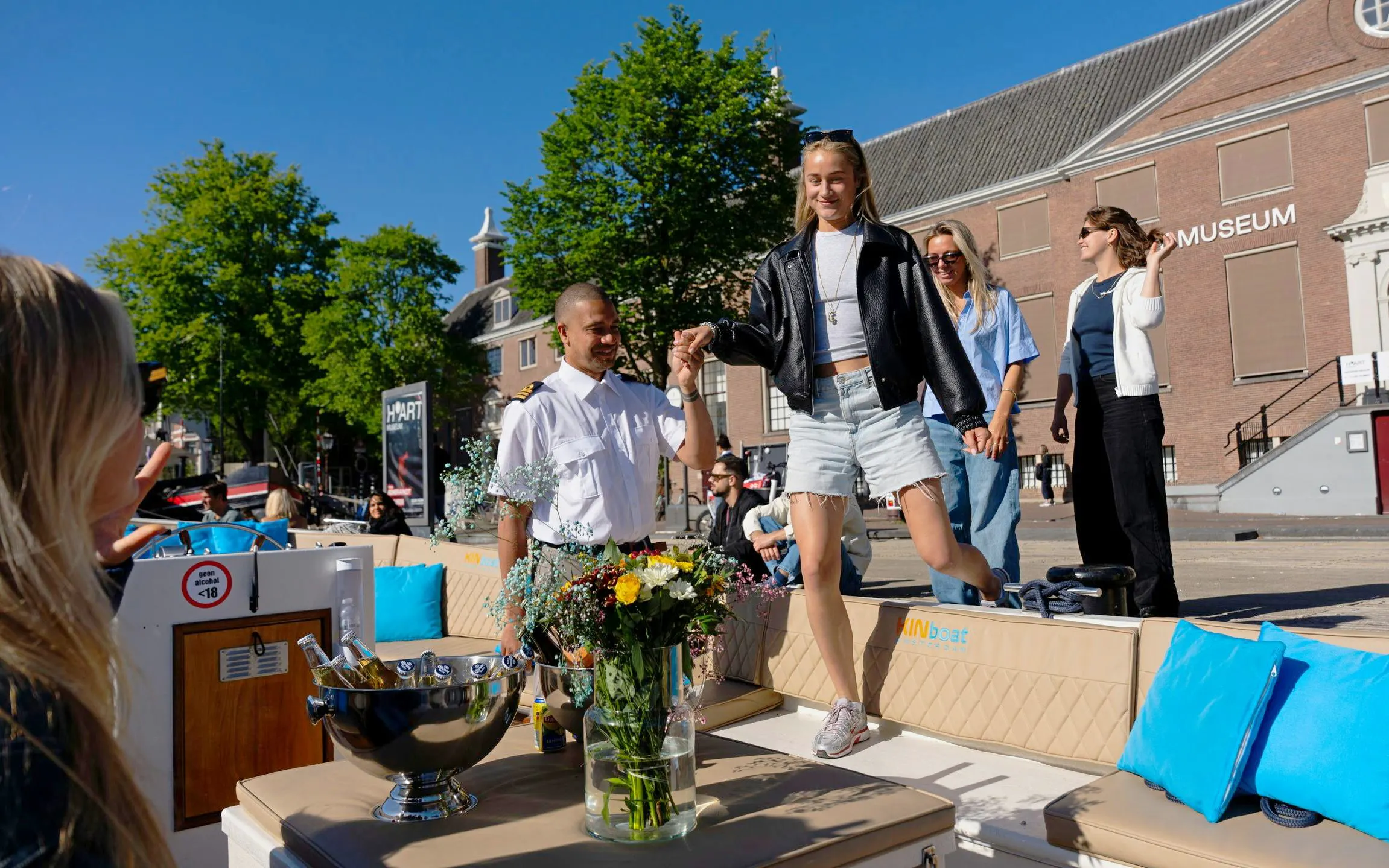 Tourists boarding an open electric boat for an Amsterdam sunset canal cruise near a museum.