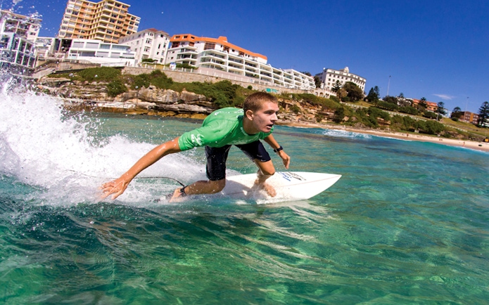Surfer riding a wave at Bondi Beach with coastal buildings in the background.
