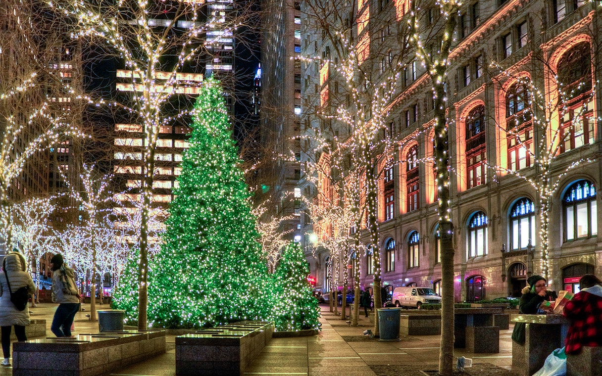 Holiday lights illuminate city street with decorated trees and buildings.