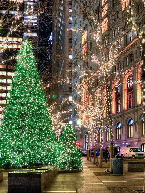 Holiday lights illuminate city street with decorated trees and buildings.