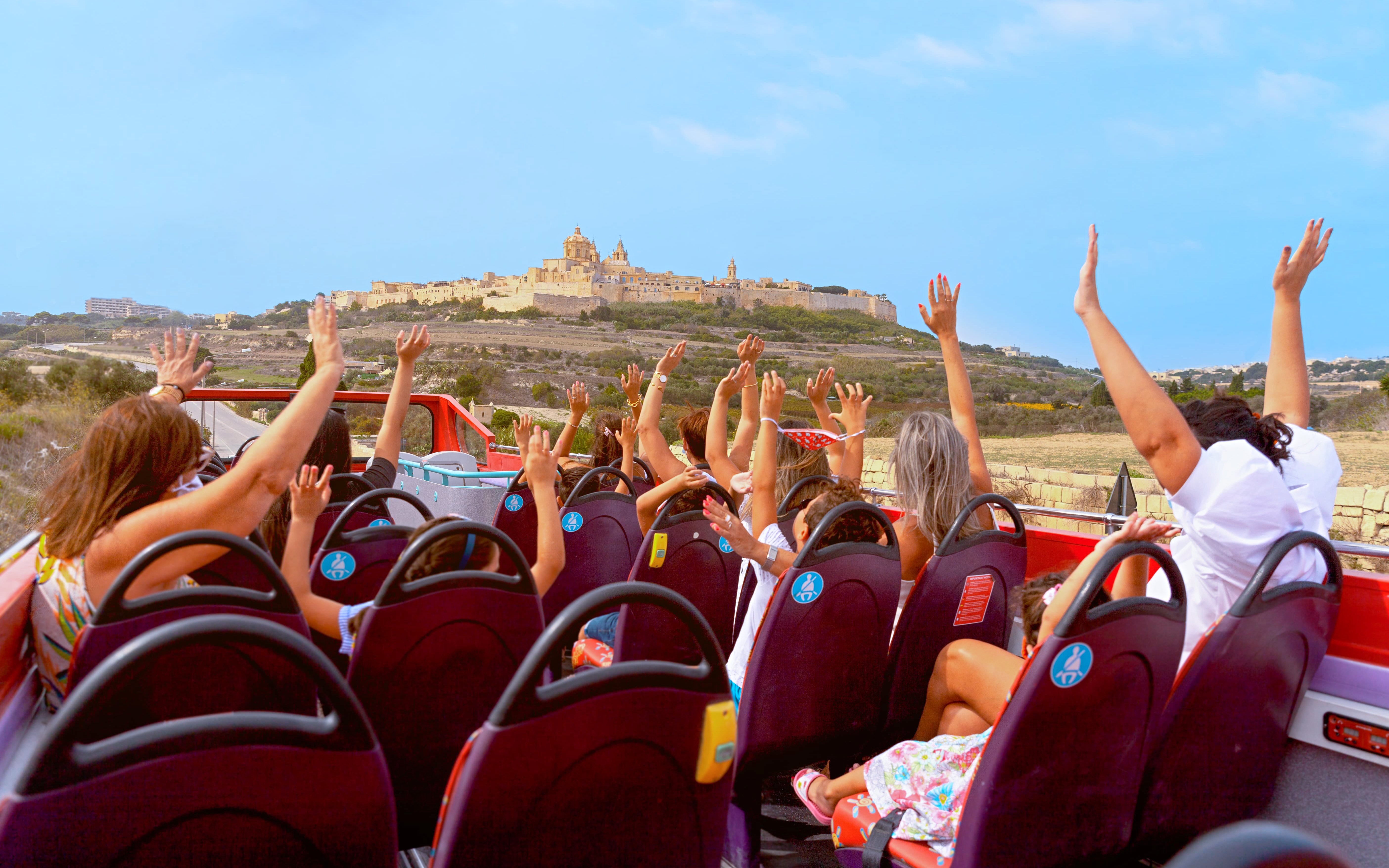 Open-top bus tour with passengers raising hands, approaching Mdina, Malta.