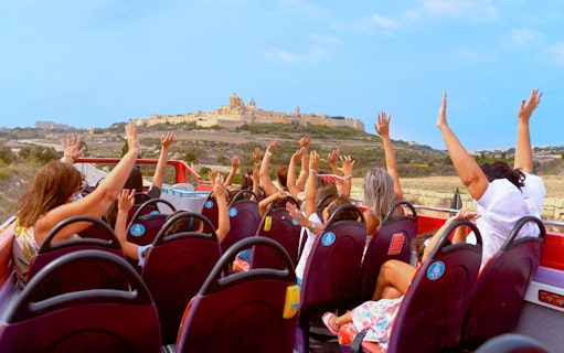 Open-top bus tour with passengers raising hands, approaching Mdina, Malta.