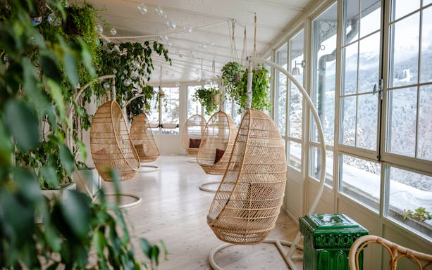 Hanging wicker chairs in a sunlit room with plants at QC Terme Bagni Nuovi di Bormio.