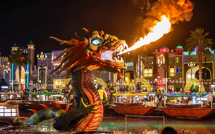Fire-breathing dragon sculpture at Global Village Dubai with illuminated pavilions in the background.