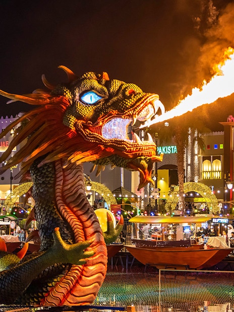 Fire-breathing dragon sculpture at Global Village Dubai with illuminated pavilions in the background.